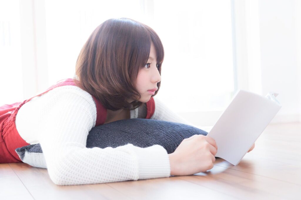 リビングの床でマンガを読みながら、奥のテレビ画面へ視線を向ける若い女性
