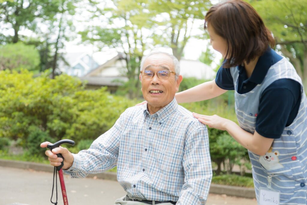 公園で杖を手に座る高齢の男性と、肩に手を添える介護士の女性