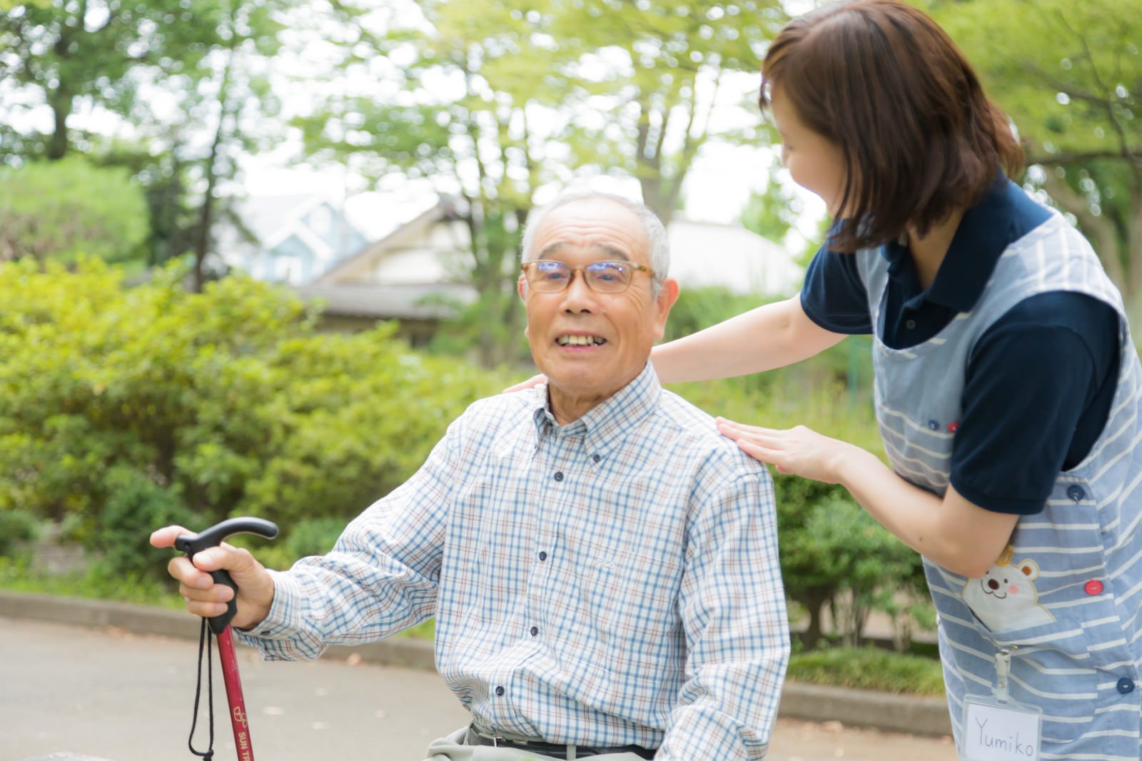 公園で杖を手に座る高齢の男性と、肩に手を添える介護士の女性
