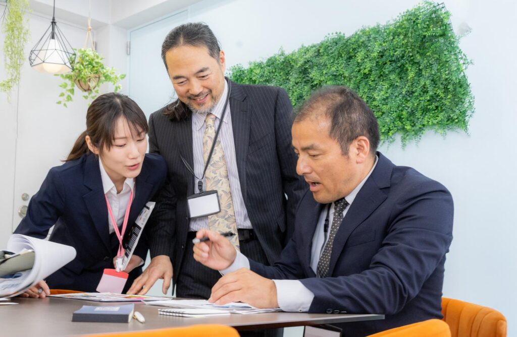 テーブルに広げた書類を指しながら、スーツ姿の女性が年配の男性2人に内容を説明している様子